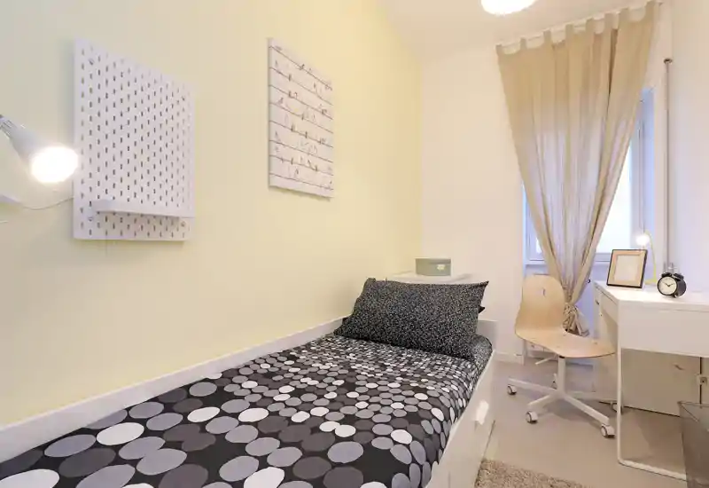 Chambre simple, compacte et lumineuse avec une couette à motifs, des étagères près du lit et un petit bureau près de la fenêtre — idéale pour un étudiant ou une personne seule.