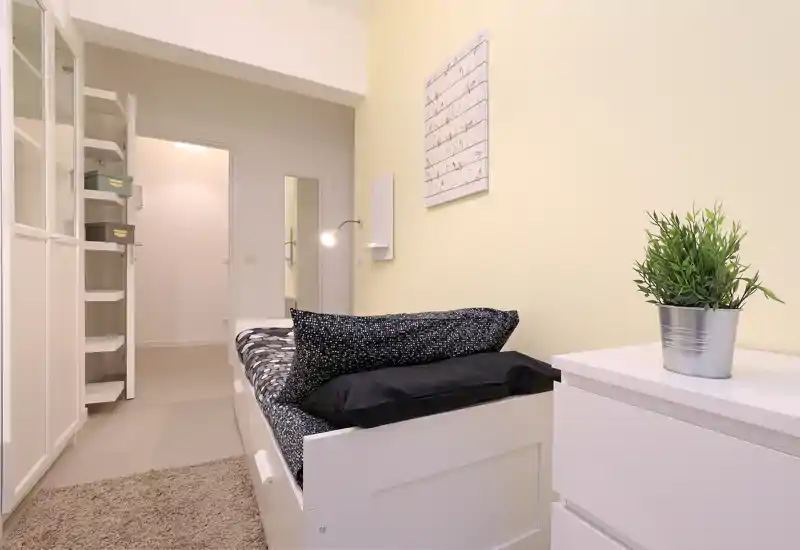 Chambre simple soignée avec un lit blanc visible, linge de lit noir et blanc, table de chevet et plante décorative. Agencement compact, lumineux et bien stylé pour un séjour confortable.