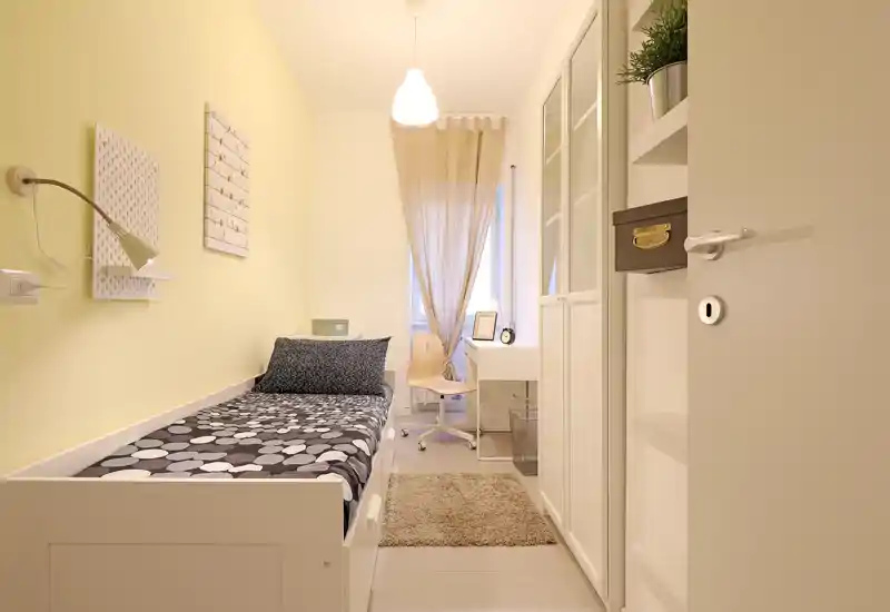 Chambre simple compacte et lumineuse avec lit blanc, literie à motifs, petit bureau près de la fenêtre et décoration aux tons chauds — idéale pour une location ou un étudiant.