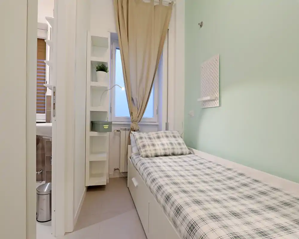 Chambre simple compacte et bien rangée avec un lit banquette en linge à carreaux, mur vert menthe, rangements intégrés et une fenêtre avec rideau laissant passer une lumière douce.