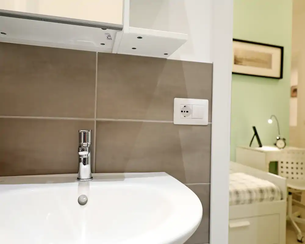Gros plan sur un lavabo de salle de bains avec robinet chromé et crédence carrelée ; une chambre est visible en arrière-plan par la porte.