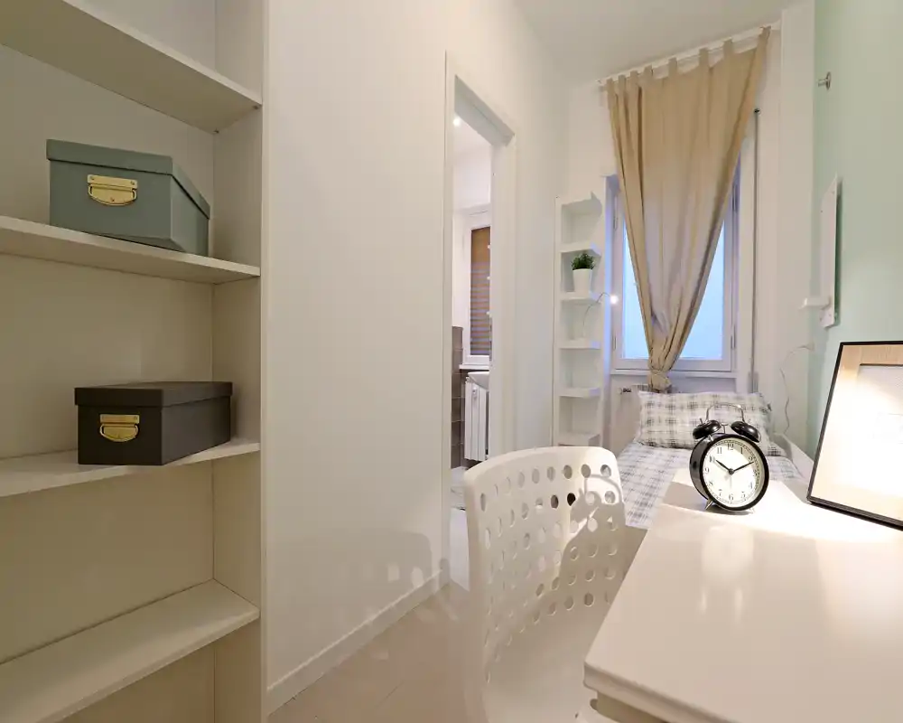 Chambre simple compacte et lumineuse avec bureau de chevet et étagères. Le lit bien fait avec parure à carreaux, la fenêtre avec rideau et la décoration minimaliste offrent un espace agréable et fonctionnel pour une personne.