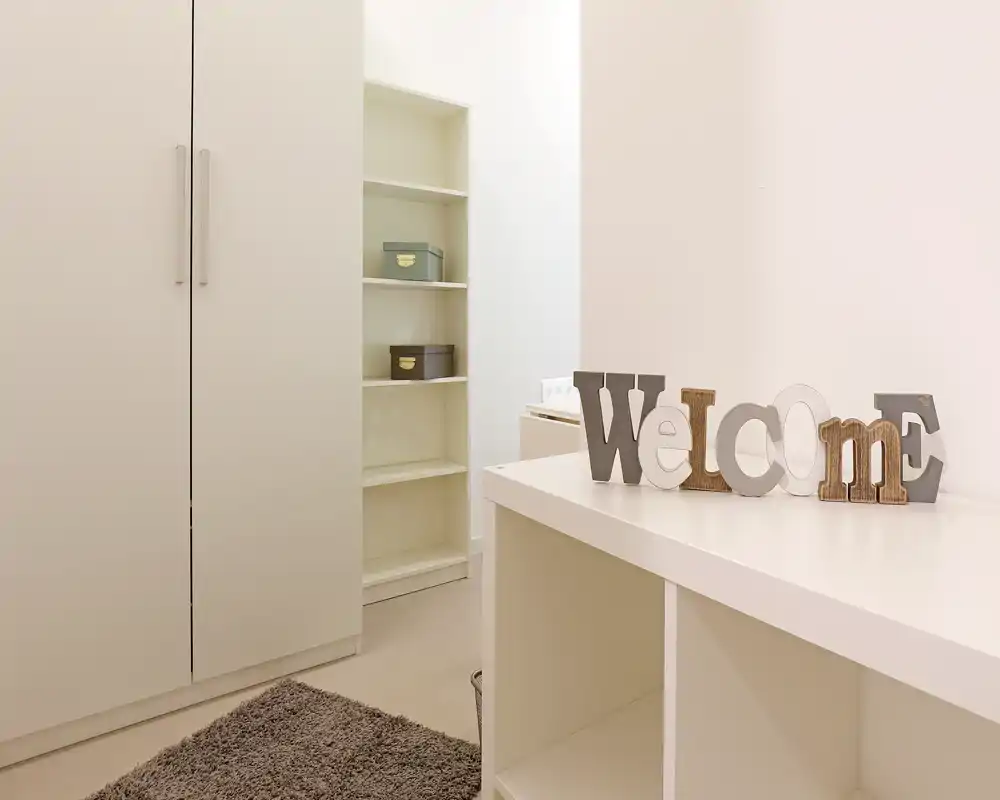 Espace de vie minimal et lumineux avec rangements intégrés, une déco "Welcome" et un tapis moelleux, parfait pour un coin entrée/salon soigné.