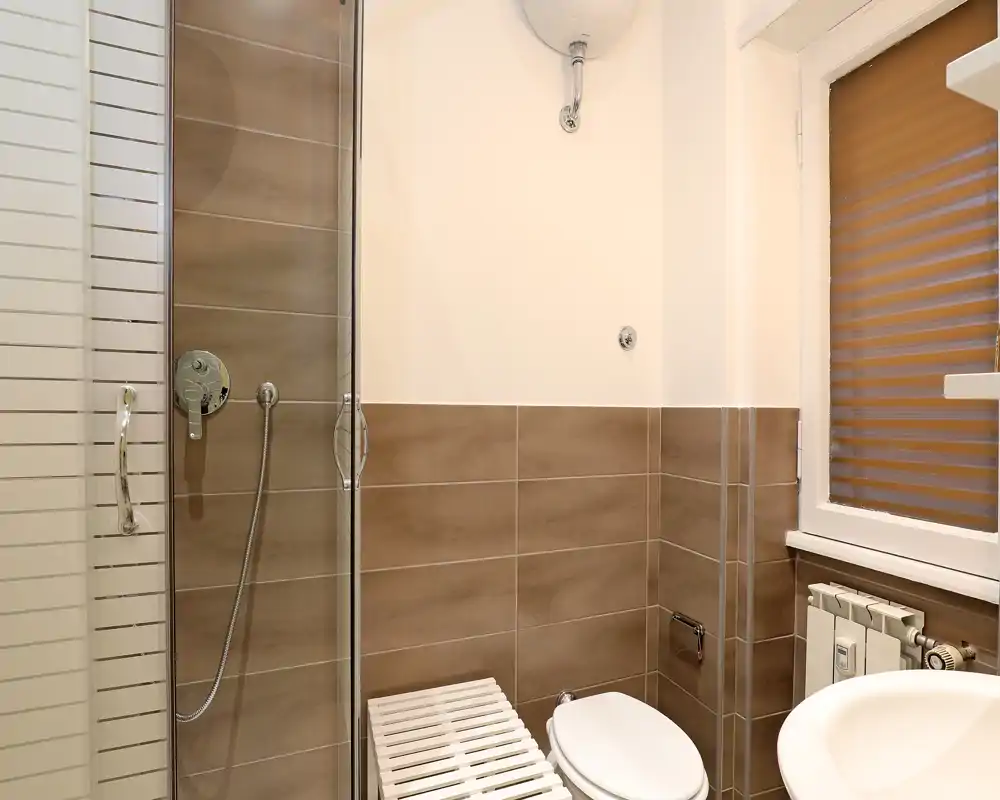 Salle de bains compacte et moderne avec cabine de douche en verre, équipements muraux et carreaux bruns neutres — lumineuse et propre.
