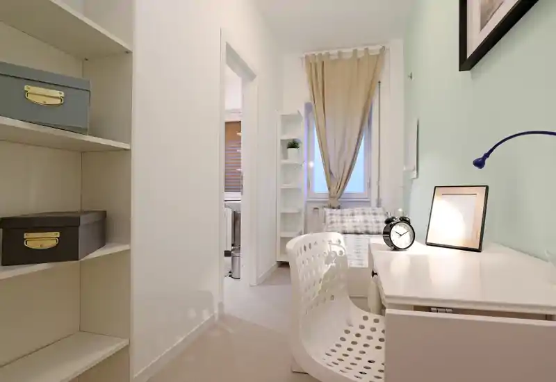 Petite chambre lumineuse avec lit simple, bureau et étagères. Des tons neutres et un rideau doux créent une ambiance calme et cosy, parfaite pour une annonce locative.