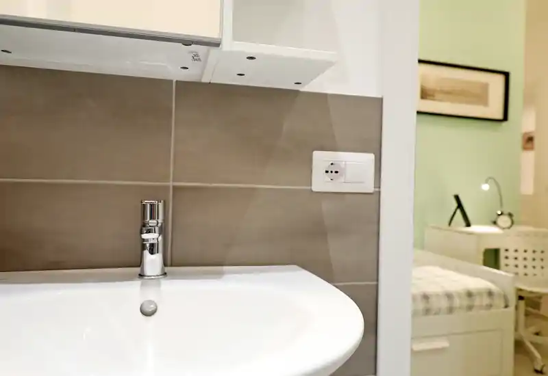 Gros plan sur un lavabo de salle de bains avec robinet chromé et crédence carrelée ; une chambre est visible en arrière-plan par la porte.