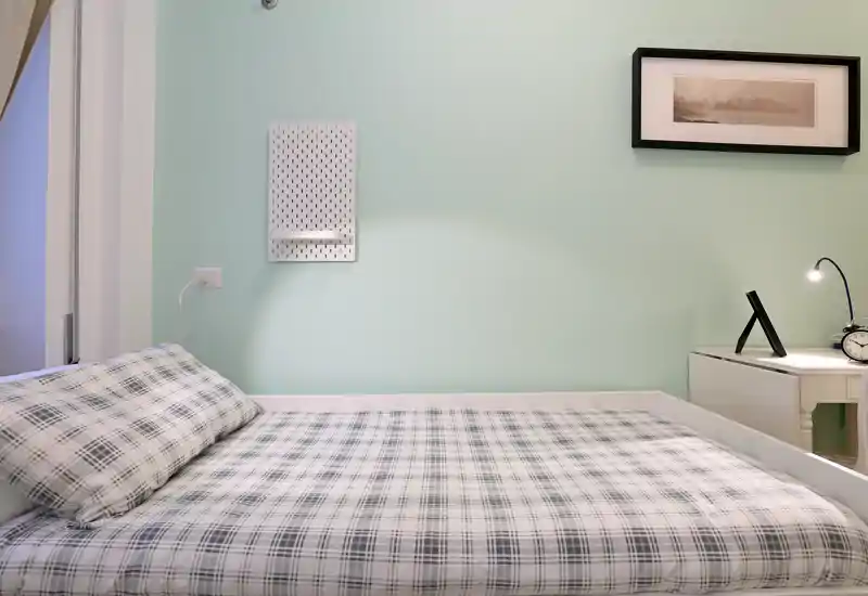 Chambre lumineuse et bien rangée avec un lit au premier plan, linge à carreaux et une table de chevet avec lampe et réveil.