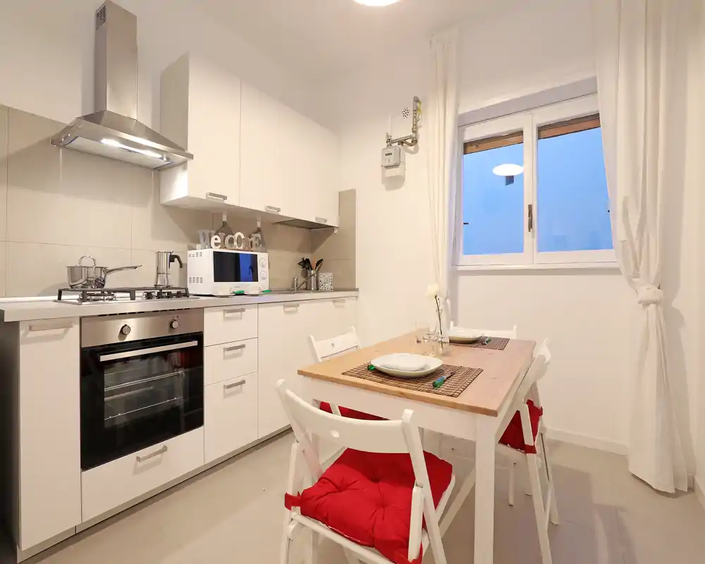 Modern compact kitchen with white cabinetry, stainless-steel appliances and a small wooden dining table set for two; bright, clean and efficiently arranged for short stays.