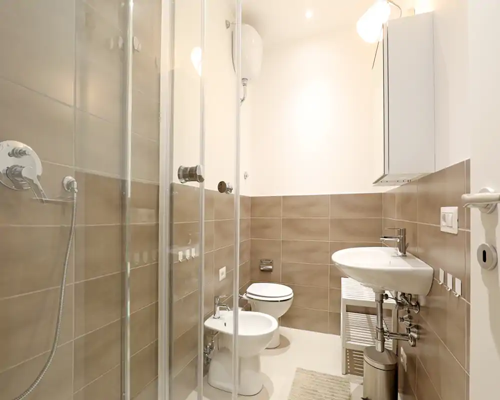 Bright, modern bathroom with a glass shower enclosure, bidet, toilet and wall-mounted sink; neutral beige tiles and clean fixtures create a fresh, functional space.