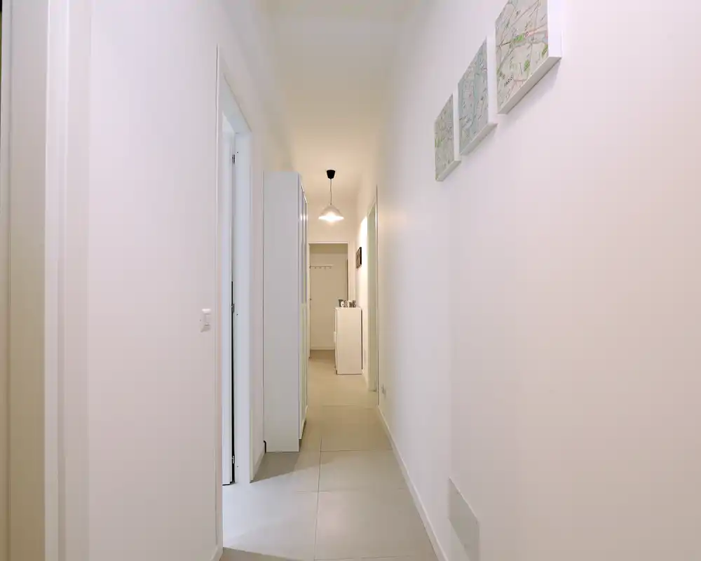 Bright, minimalist entrance corridor with tiled floor, wall décor and overhead pendant lighting — clean and welcoming access to the apartment.