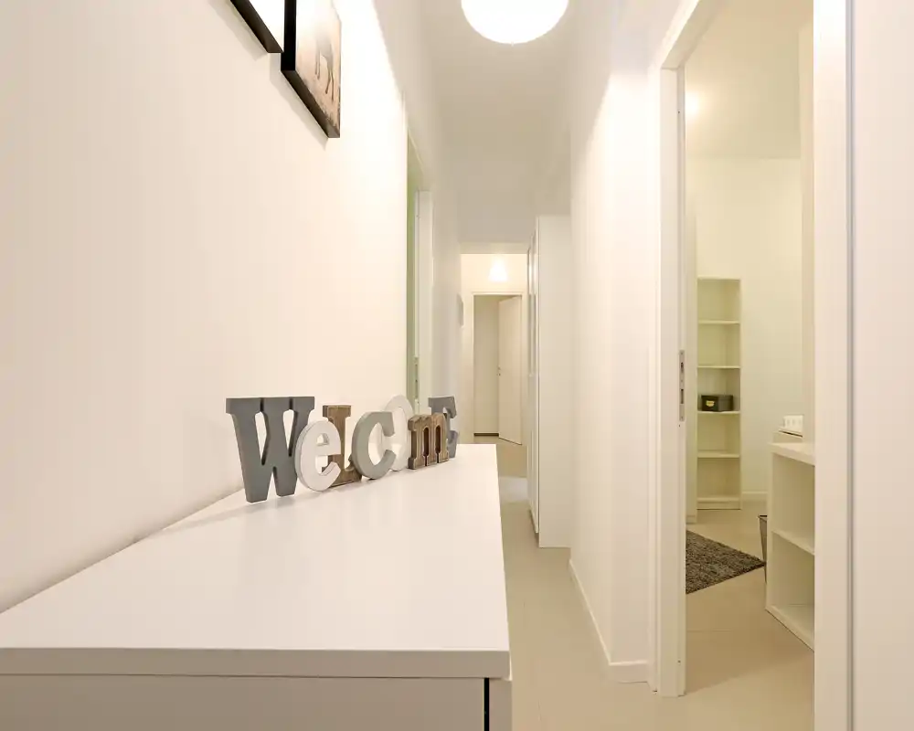 Bright, minimal hallway with a console table and decorative 'Welcome' letters; neutral tones and recessed doors lead to adjacent rooms.