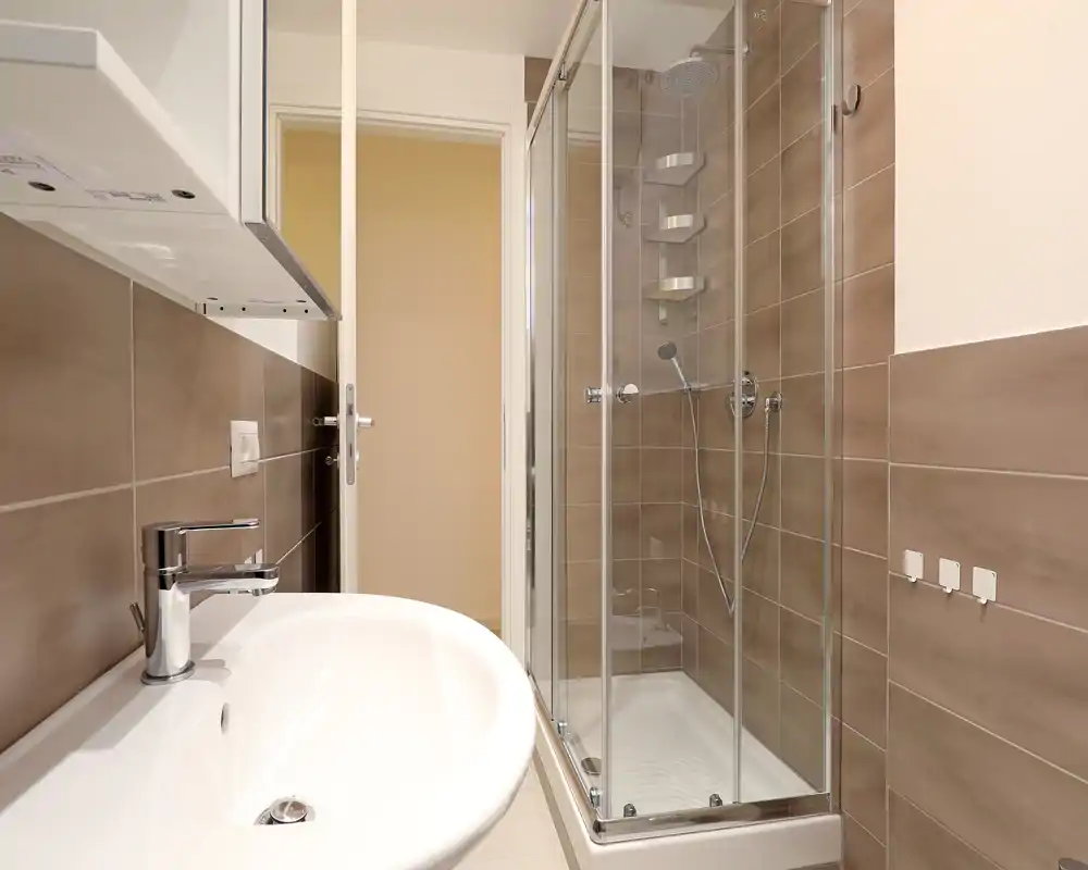 Modern compact bathroom with glass-enclosed shower, wall-mounted fixtures and neutral beige tiles — clean and well lit.