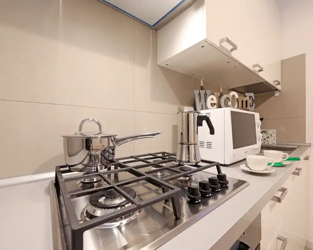Modern compact kitchen corner with a stainless-steel gas hob in the foreground, a moka pot, microwave and small decorative details on the counter.