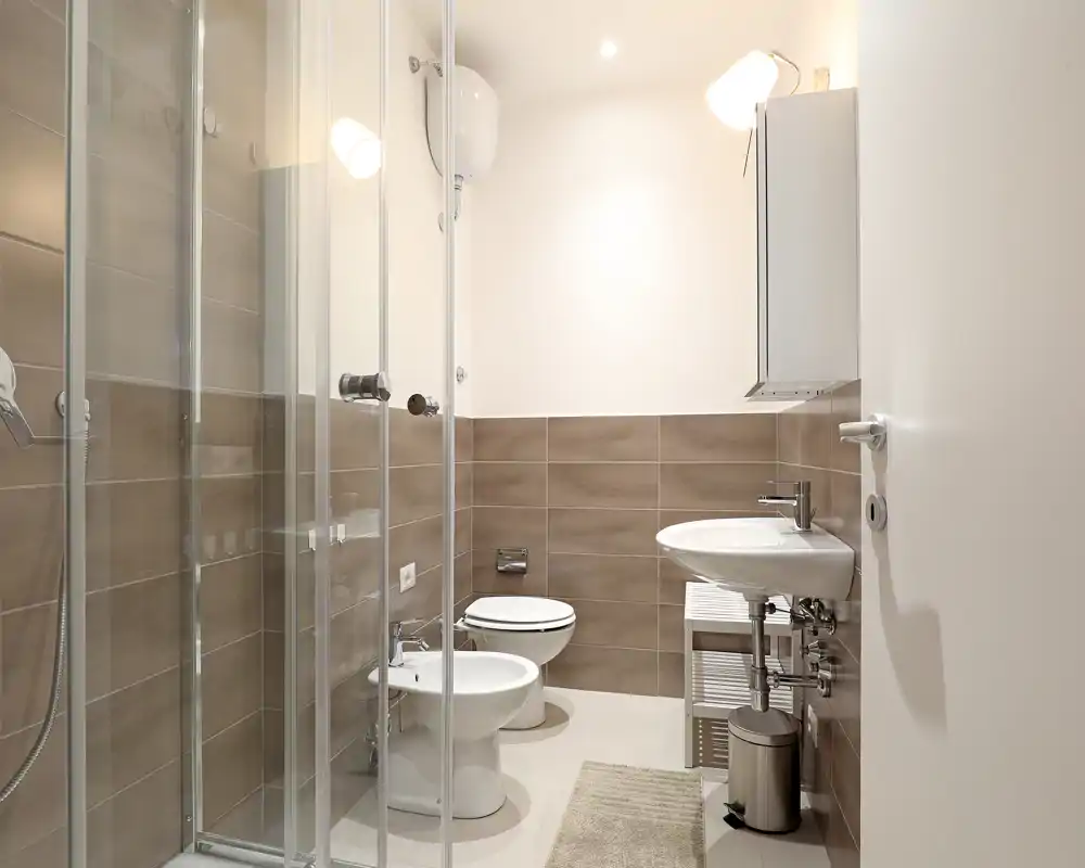 Clean, modern bathroom with a glass-enclosed shower, bidet, toilet and wall-mounted sink in neutral tones; bright, well-lit and neatly arranged.