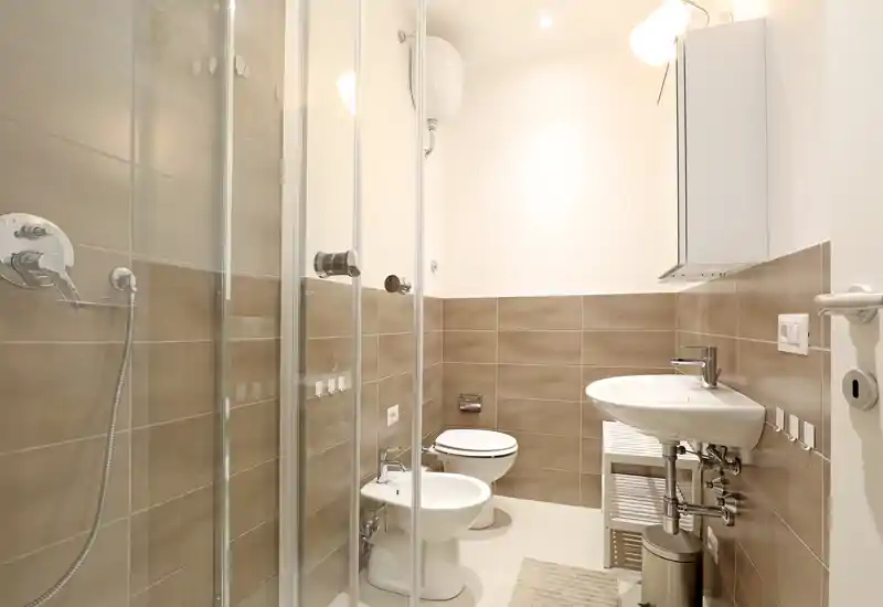 Salle de bains moderne et lumineuse avec cabine de douche en verre, bidet, WC et lavabo mural; carreaux beiges neutres et installations propres pour un espace fonctionnel.