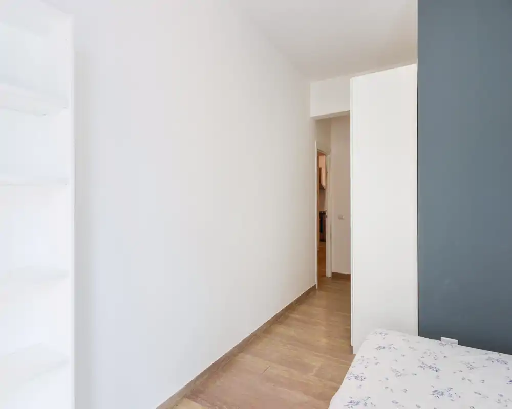 Coin de chambre avec un lit partiellement visible, murs clairs et sol en bois ; lumineux, minimal et prêt à être aménagé.