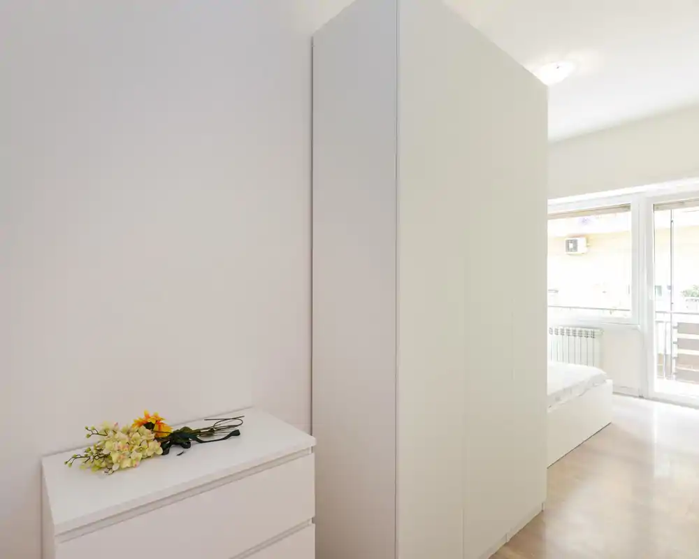 Coin chambre lumineux et minimaliste avec une armoire blanche, une commode ornée de fleurs et un lit partiellement visible près d'une grande fenêtre laissant entrer beaucoup de lumière naturelle.
