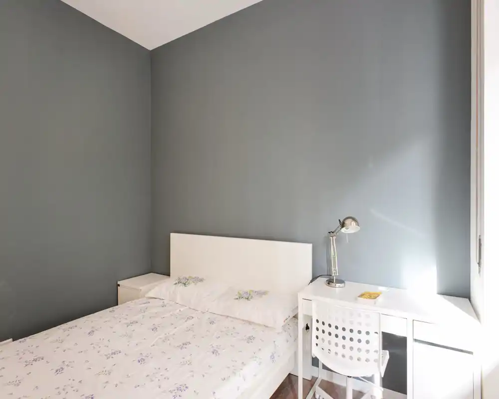Chambre lumineuse et minimaliste avec lit blanc, tables de chevet et bureau près de la fenêtre — linge doux et lampe de lecture pour une ambiance calme.