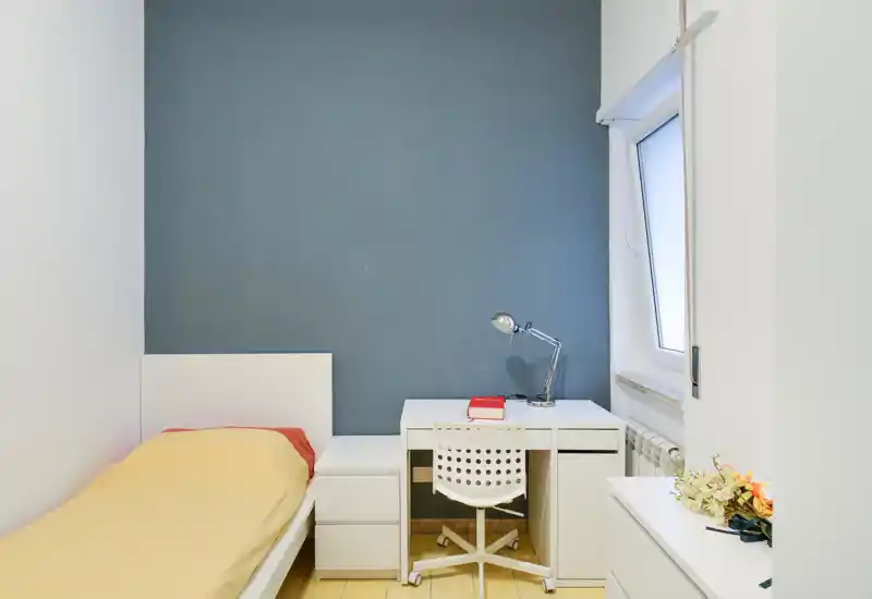 Camera singola luminosa e semplice con letto giallo, mobili bianchi e una scrivania compatta accanto alla finestra. Stile minimal pulito e parete blu che dona carattere, adatta per affitti brevi o studenti.