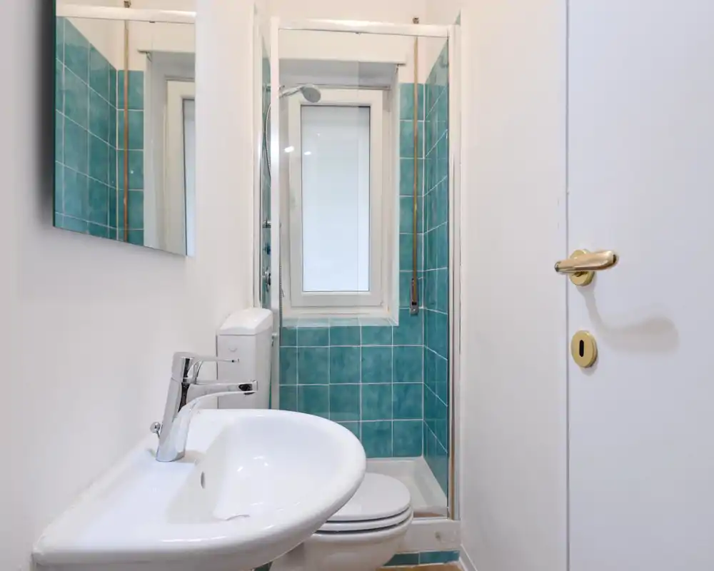 Bagno compatto con doccia piastrellata turchese, lavabo e WC bianchi; spazio luminoso, pulito e ben illuminato, ideale per un piccolo affitto.
