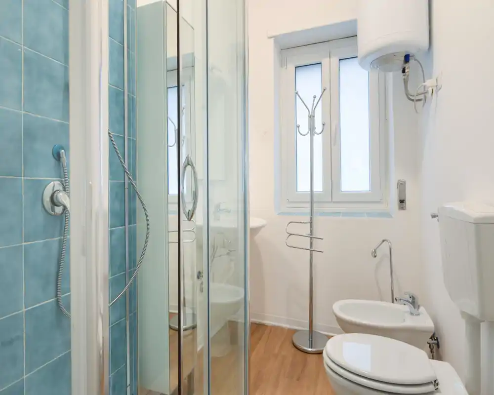 Bagno compatto e luminoso con box doccia in vetro, parete piastrellata blu, bidet e wc; pulito, ben illuminato, adatto per annunci di affitto.