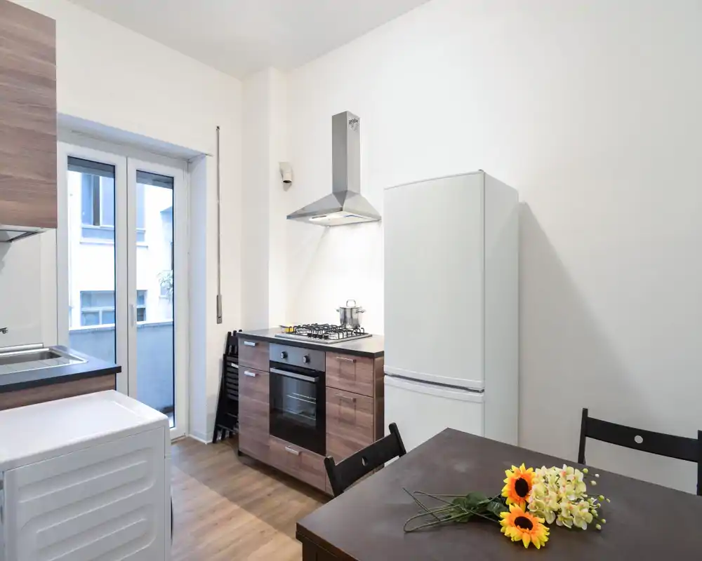 Cucina compatta e moderna con mobili effetto legno, piano cottura a gas e cappa in acciaio; finestra luminosa, frigorifero visibile e tavolo da pranzo con fiori che aggiungono un tocco accogliente.