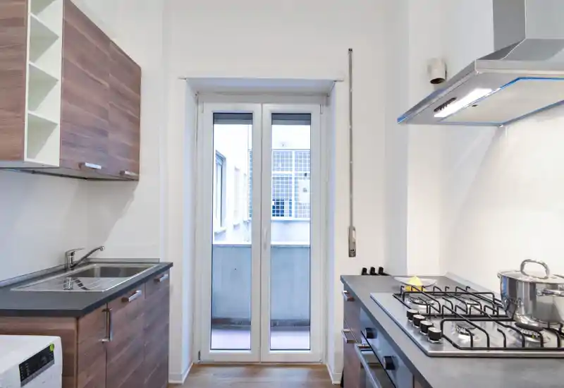 Cucina moderna e luminosa con mobili effetto legno, lavello in acciaio e piano cottura a gas; luce naturale grazie alla porta finestra verso un piccolo balcone.