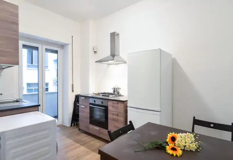 Cucina compatta e moderna con mobili effetto legno, piano cottura a gas e cappa in acciaio; finestra luminosa, frigorifero visibile e tavolo da pranzo con fiori che aggiungono un tocco accogliente.