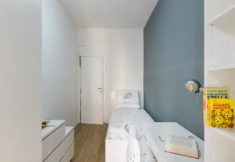 Dormitorio individual compacto con cama blanca ordenada, escritorio y estanterías; pared azul de acento y luz natural crean un ambiente minimalista y cuidado.