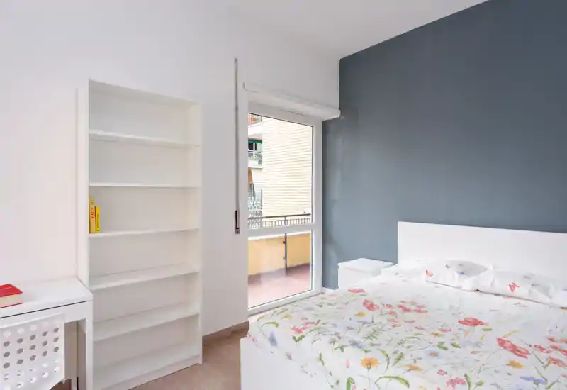 Dormitorio luminoso y minimalista con cama blanca y ropa de cama floral, mesita de noche y una estantería blanca junto a una gran ventana con balcón.