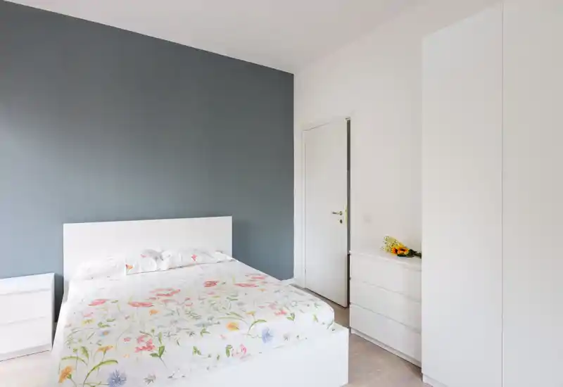 Dormitorio moderno y luminoso con cama blanca y edredón floral frente a una pared azul tenue. Líneas limpias, luz natural y decoración mínima crean una atmósfera tranquila y acogedora ideal para anuncios.