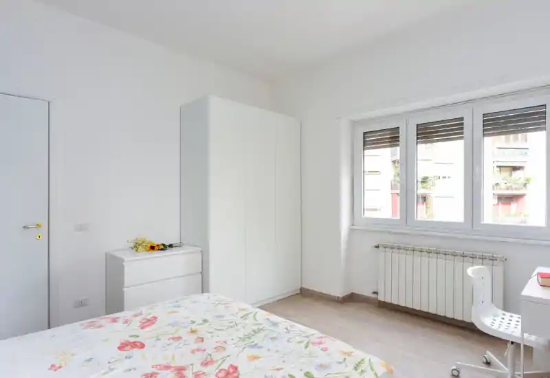 Dormitorio luminoso y minimalista con cama con colcha floral, armario blanco y una gran ventana que aporta luz natural abundante.