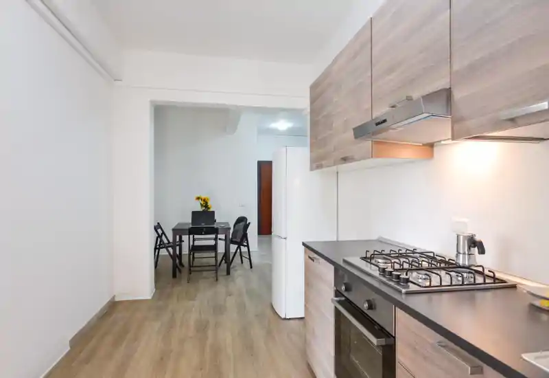 Cocina abierta moderna con muebles efecto madera, cocina de gas y pequeño comedor; luminosa y limpia, perfecta para apartamentos compactos.