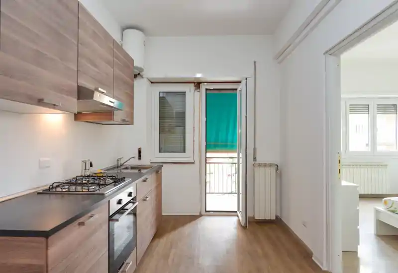 Cocina compacta moderna con muebles efecto madera, horno empotrado y placa de gas, luz natural desde la puerta/balcón y suelo laminado bien cuidado.