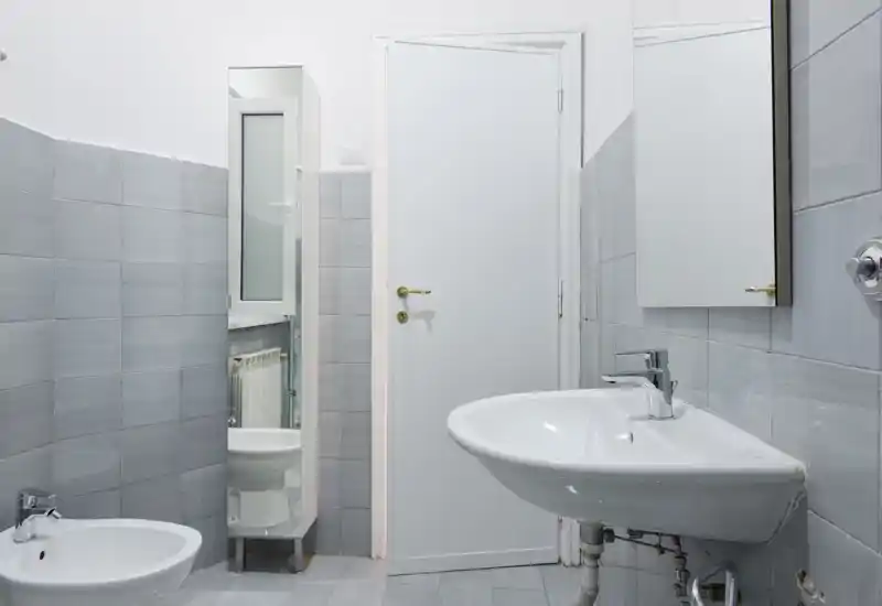 Baño compacto y limpio con lavabo suspendido, bidé, armario con espejo y azulejos grises neutras, bien iluminado y de estilo sencillo.