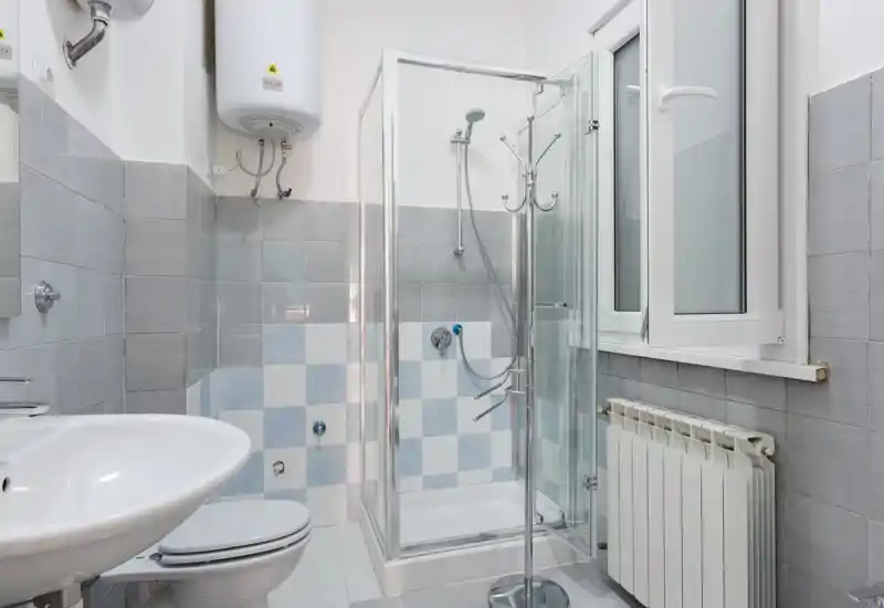 Salle de bains moderne et propre avec cabine de douche en verre, chauffe-eau mural, lavabo et WC ; carreaux gris et bleus pour un rendu frais et neutre.