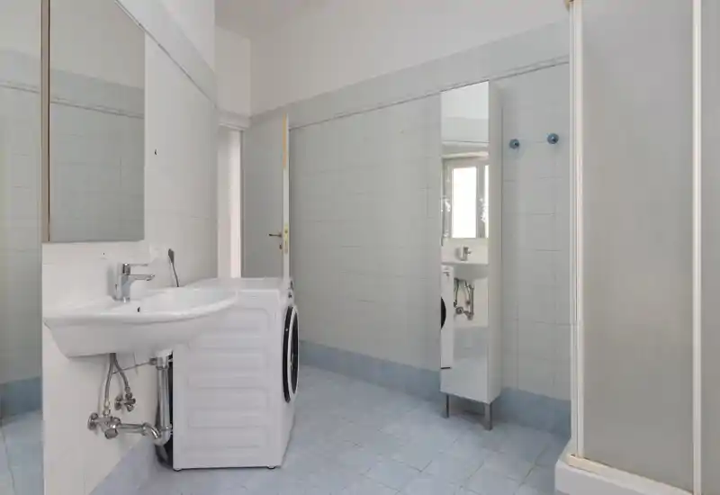Salle de bains carrelée et lumineuse avec lavabo, miroir, lave-linge et cabine de douche ; style neutre et propre avec une bonne lumière naturelle.
