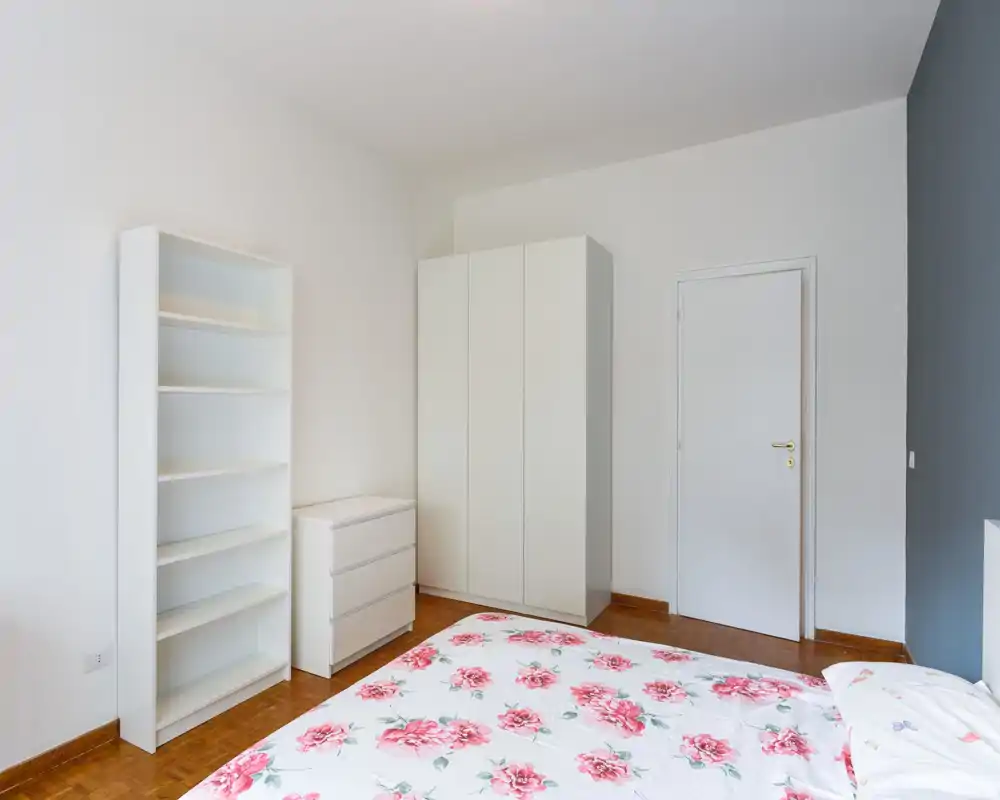 Camera da letto luminosa e minimal con copriletto floreale, armadio e scaffali bianchi che offrono spazio e un’atmosfera neutra e ordinata.