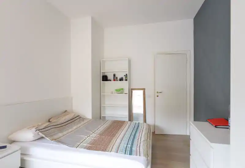 Camera da letto minimale e luminosa con letto matrimoniale in primo piano, copriletto a righe, una libreria bianca e una cassettiera; ambiente pulito e moderno.