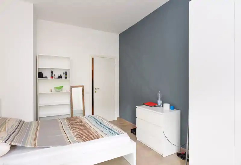 Camera da letto luminosa e minimal con letto ben visibile, comò bianco e mensole; toni neutri e parete accentuata scura per un'atmosfera moderna e tranquilla.