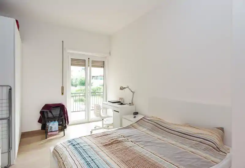Camera da letto luminosa e minimale con letto matrimoniale, comodino e scrittoio vicino alla porta del balcone che lascia entrare luce naturale.