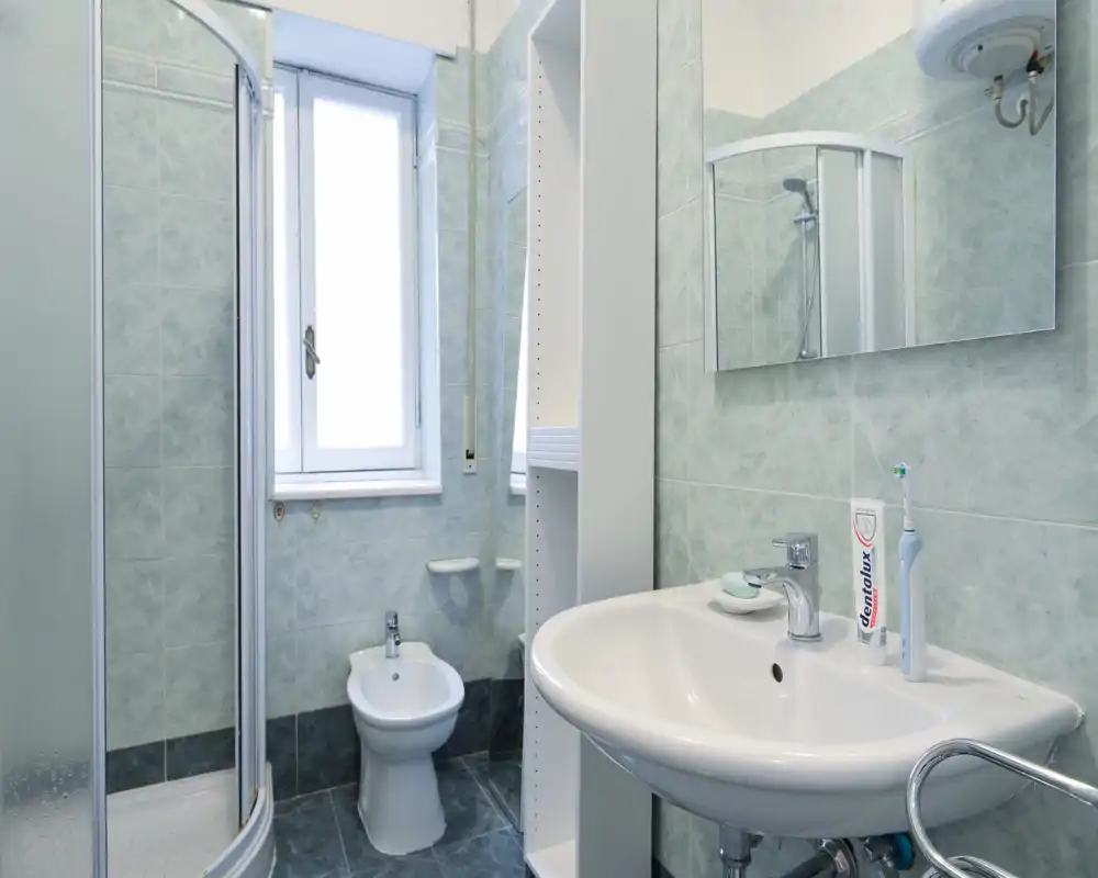 Bagno compatto e pulito con doccia ad angolo, bidet e lavabo a muro; piastrelle verde chiaro e specchio rendono l'ambiente luminoso e ordinato.