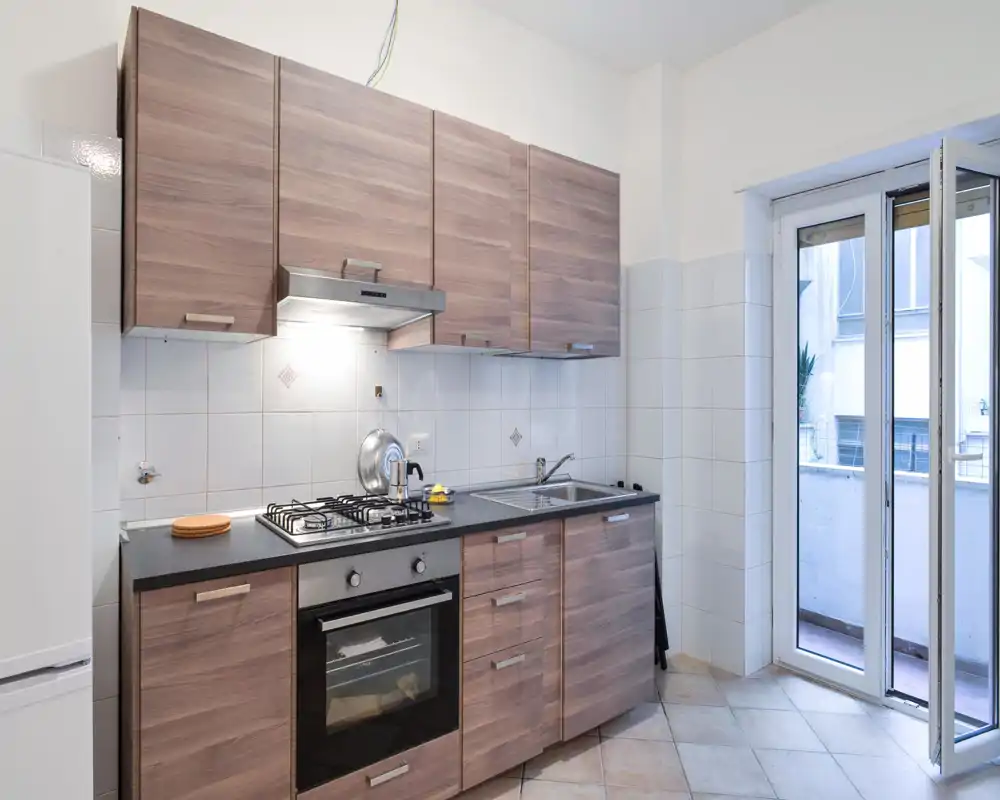 Cucina compatta e moderna con ante effetto legno, forno e piano cottura a gas integrati, lavello in acciaio e accesso diretto a un piccolo balcone—luminoso, pulito e funzionale.
