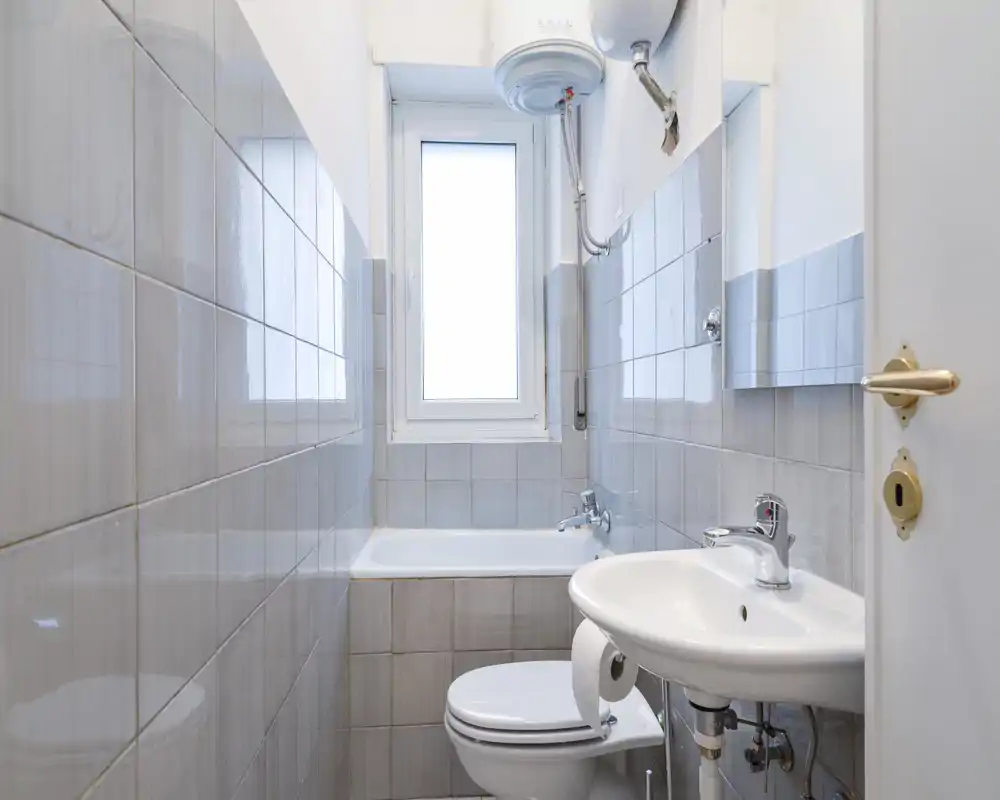 Bagno compatto con pareti piastrellate grigie, vasca sotto la finestra centrale, scaldabagno a parete e lavabo con rubinetteria cromata; spazio luminoso e funzionale ideale per un appartamento piccolo.