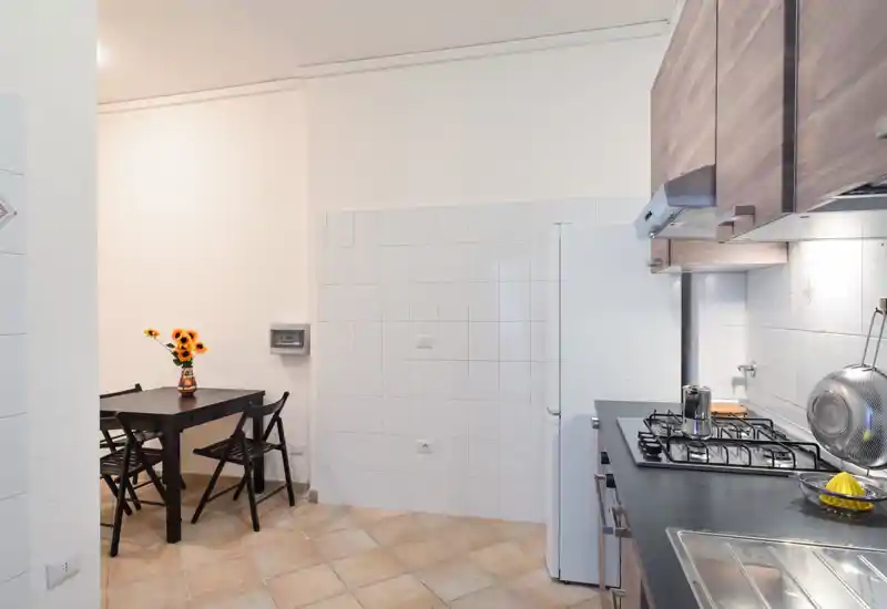 Cucina compatta e ben illuminata con piano cottura a gas, lavello e un piccolo tavolo da pranzo decorato con un vaso di girasoli: pratica e accogliente per un appartamento in città.