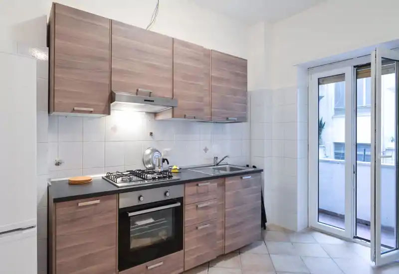 Cucina compatta e moderna con ante effetto legno, forno e piano cottura a gas integrati, lavello in acciaio e accesso diretto a un piccolo balcone—luminoso, pulito e funzionale.