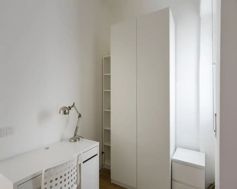 Pequeña habitación luminosa con escritorio blanco, silla, lámpara y armario alto; estilo minimalista neutro y buena luz natural.