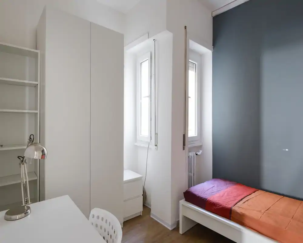 Dormitorio pequeño y luminoso con cama individual, mobiliario blanco y una lámpara de escritorio. La luz natural desde las ventanas estrechas y la pared gris crean un espacio sencillo y funcional, ideal para un estudiante o una persona sola.