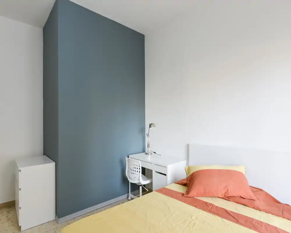 Dormitorio sencillo y luminoso con cama visible, escritorio y mesita; pared de acento azul y muebles blancos que crean un espacio minimalista y bien iluminado.