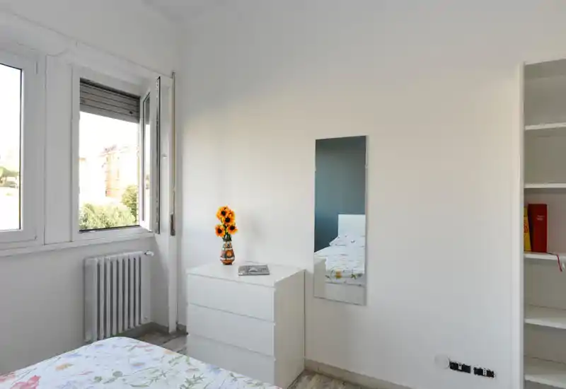 Rincón de dormitorio luminoso y minimalista con cama visible, cómoda blanca, espejo y un jarrón con girasoles; la luz natural de la ventana abierta aporta una sensación fresca y acogedora.